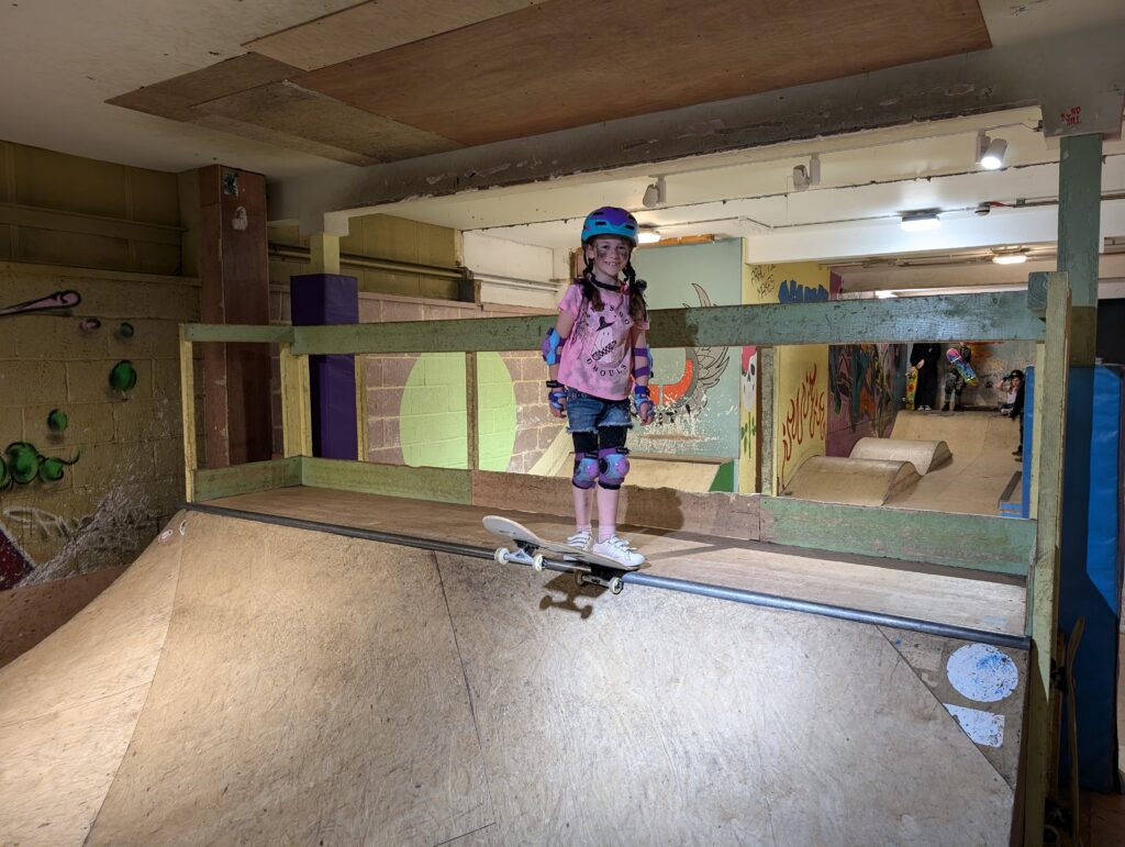 A young girl wearing a helmet and protective pads stands with her skateboard at the top of a ramp in an indoor skatepark, smiling confidently before her run.