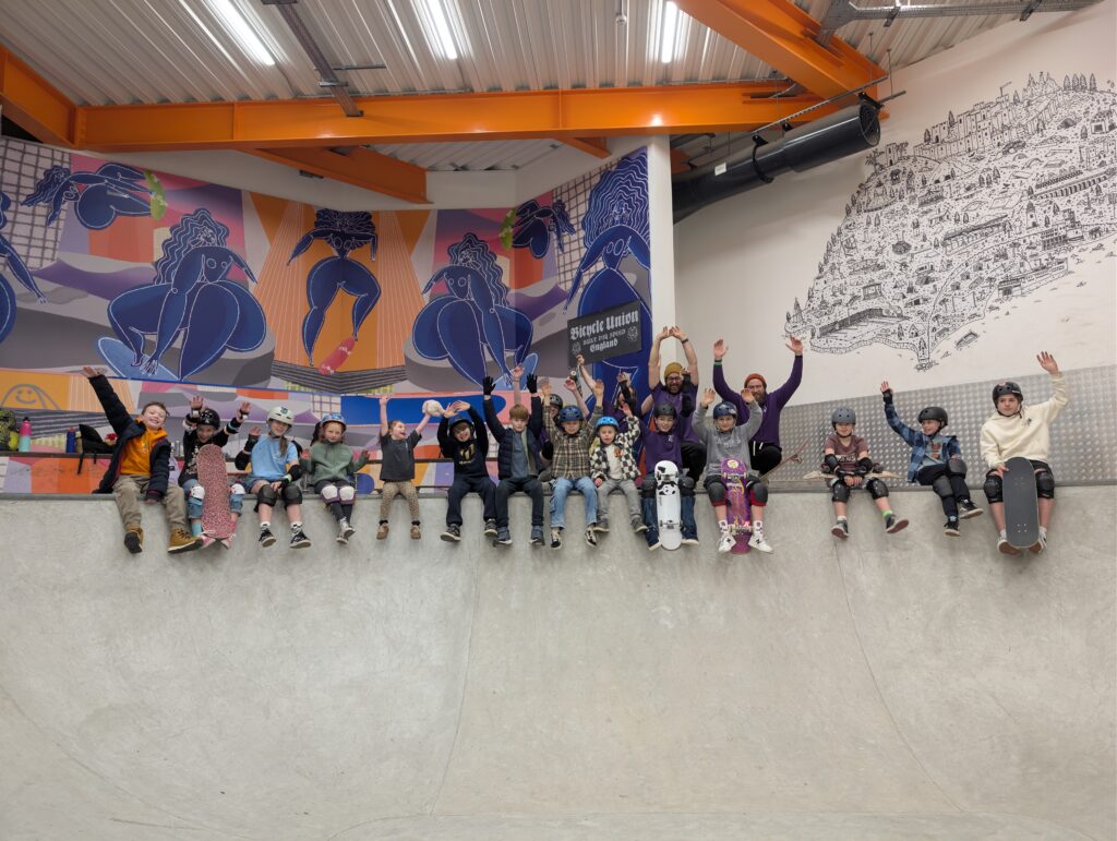 A group of children and adult instructors sit along the edge of an indoor skatepark bowl, smiling and raising their hands for a group photo. Most of the children wear helmets and protective pads, with several holding skateboards. Behind them, colourful mural artwork and large orange steel beams create a vibrant backdrop.