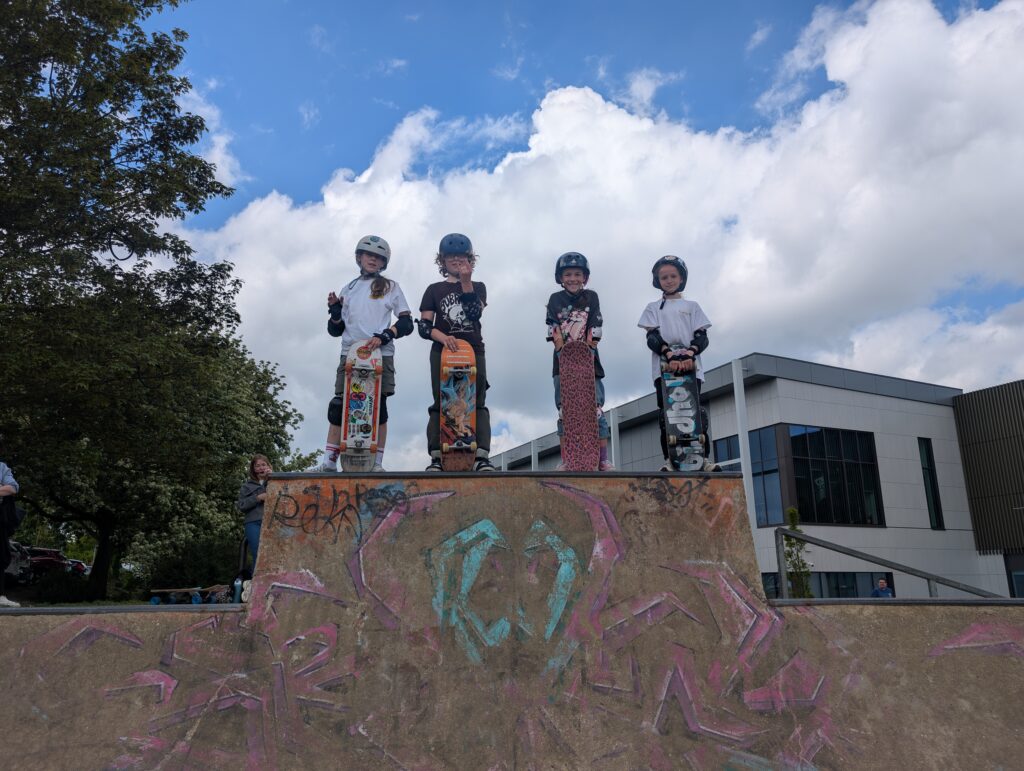 Four young skateboarders wearing helmets and protective gear stand proudly on top of a skatepark ramp holding their skateboards during a Gnarwhal Skate School session. The sky is bright with large clouds overhead, creating a fun and adventurous atmosphere.