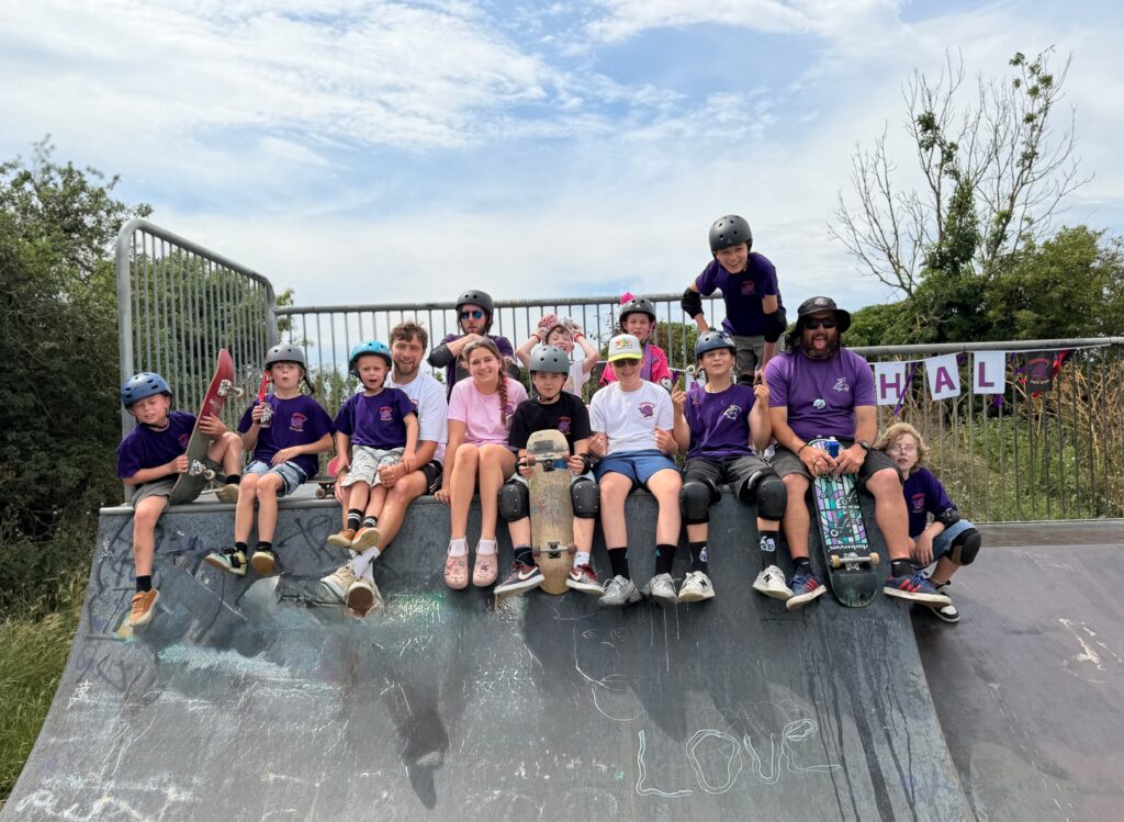 Group photo of children and coaches from Gnarwhal Skate School sitting together on a skate ramp, wearing helmets and holding skateboards during a fun community skate session in Kent.